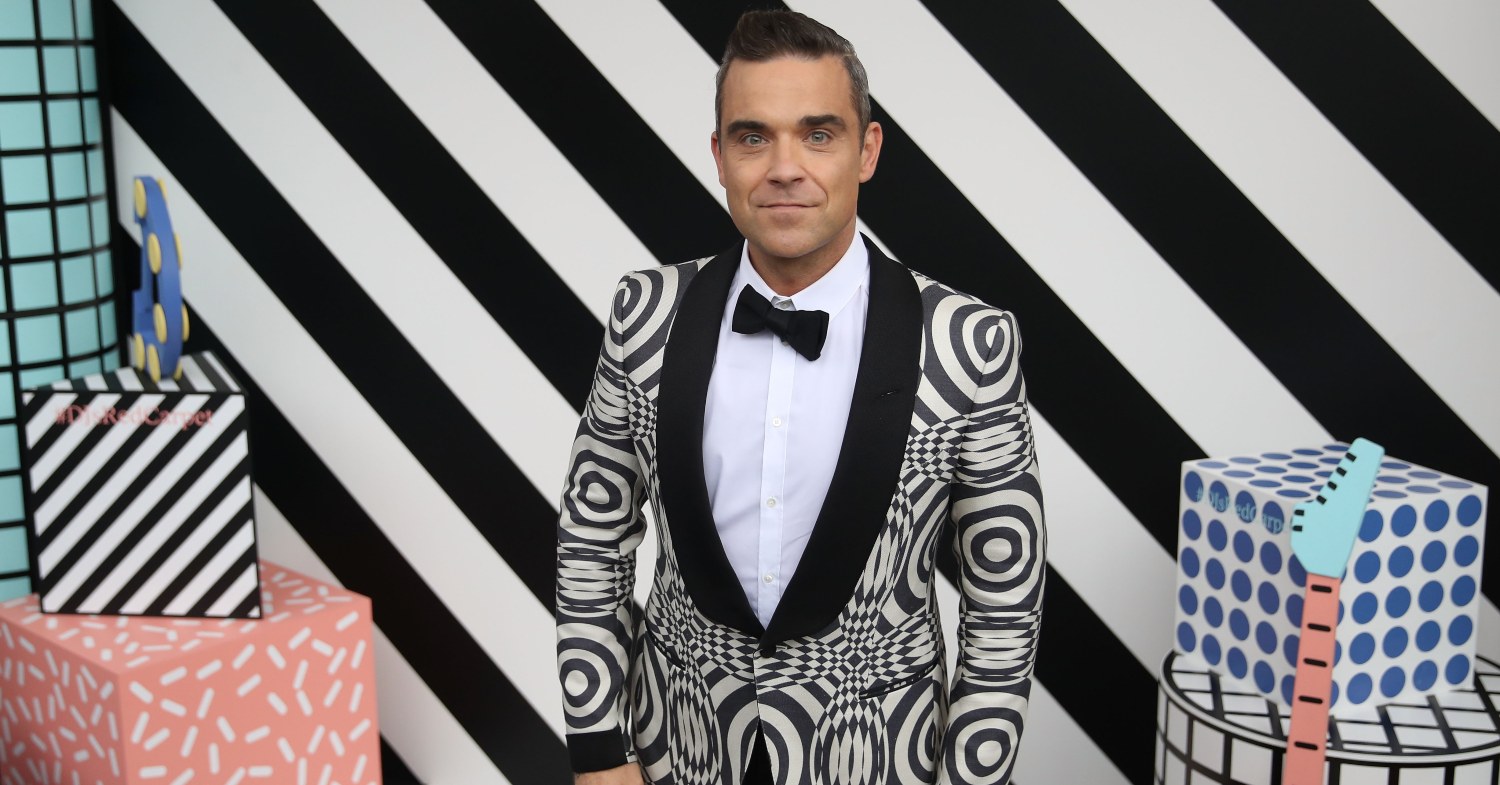 Robbie Williams stands at an event against a black and white striped background