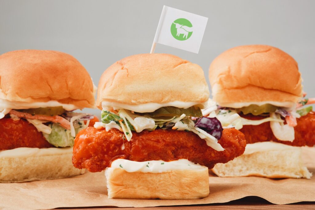 Beyond Meat's vegan chicken tenders in burger buns with pickles, salad, and wing sauce, featuring a Beyond flag and a grey background.