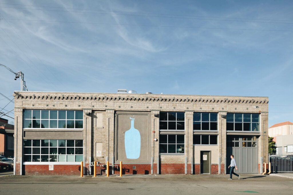 Photo of the front of a Blue Bottle Coffee shop and roastery. All the brand's branches now serve oatPhoto of the front of a Blue Bottle Coffee shop and roastery. All branches now serve oat milk. milk.
