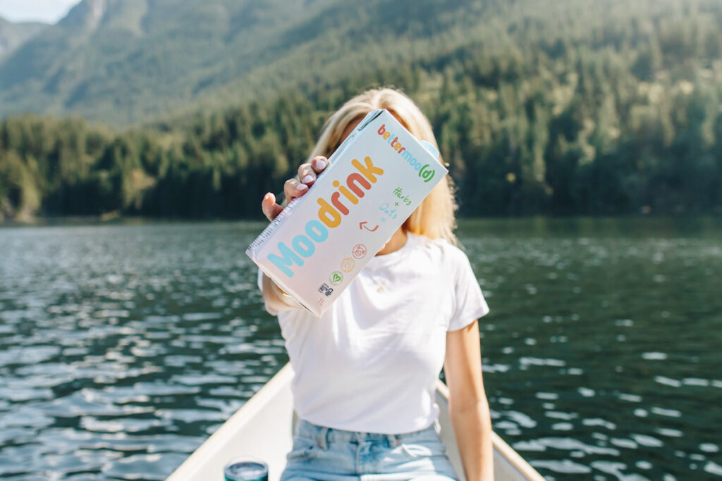 Photo shows a blond woman sat in a boat on a scenic lake holding a cartoon of Bettermood's "Moo Drink" plant-based milk up in front of her face.