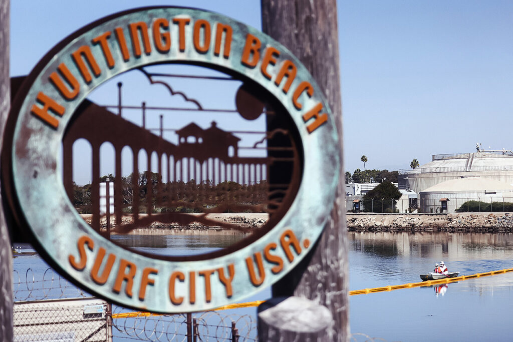 Photo shows a sign that reads: "Huntington Beach Surf City USA," with an inlet and boat in the background.
