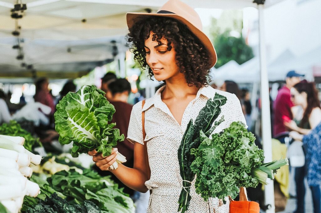Photo shows a women shopping for fresh produce at an outdoor market and is holding two different varieties of leafy greens.
