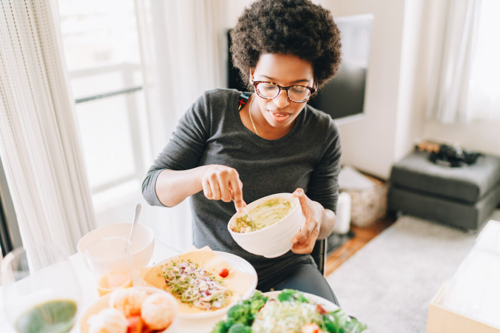 Photo shows a young woman preparing vegan food at home using salad, fruit, and vegetables. 