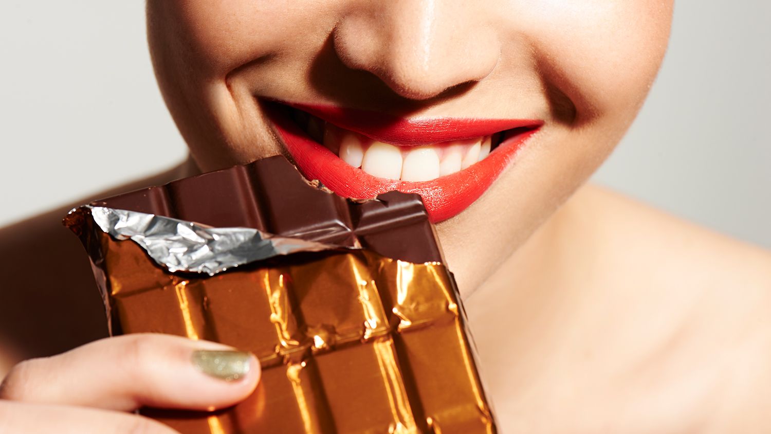 Photo showing a close-up of a person wearing red lipstick and holding a chocolate bar wrapped in gold foil