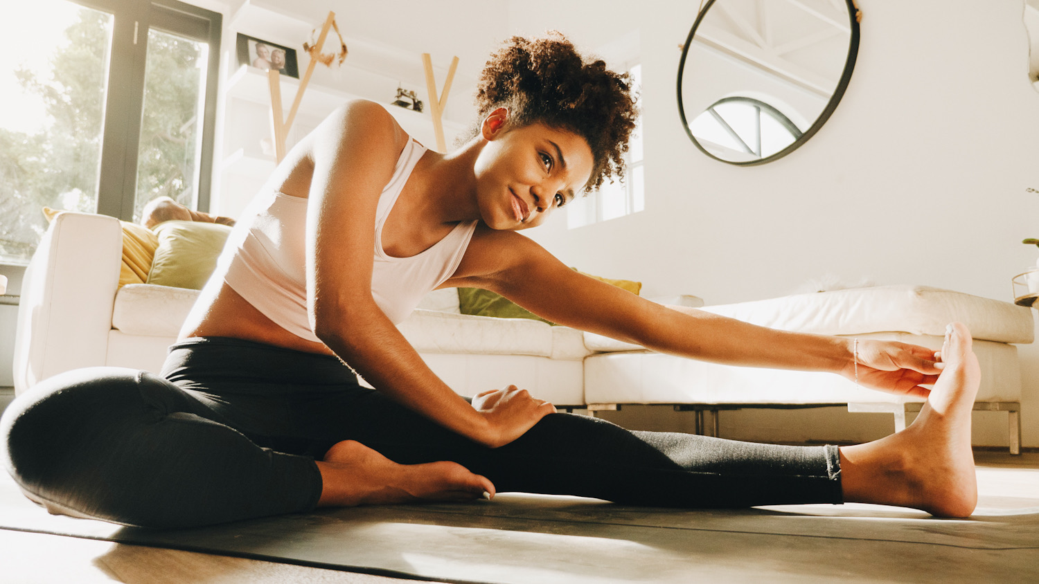 Photo shows a woman stretching on a yoga mat in a home setting. Gift the gift of self care this holiday season.