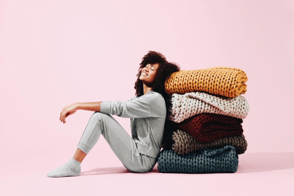 Photo shows a woman leaning against a stack of Bearaby weighted blankets.