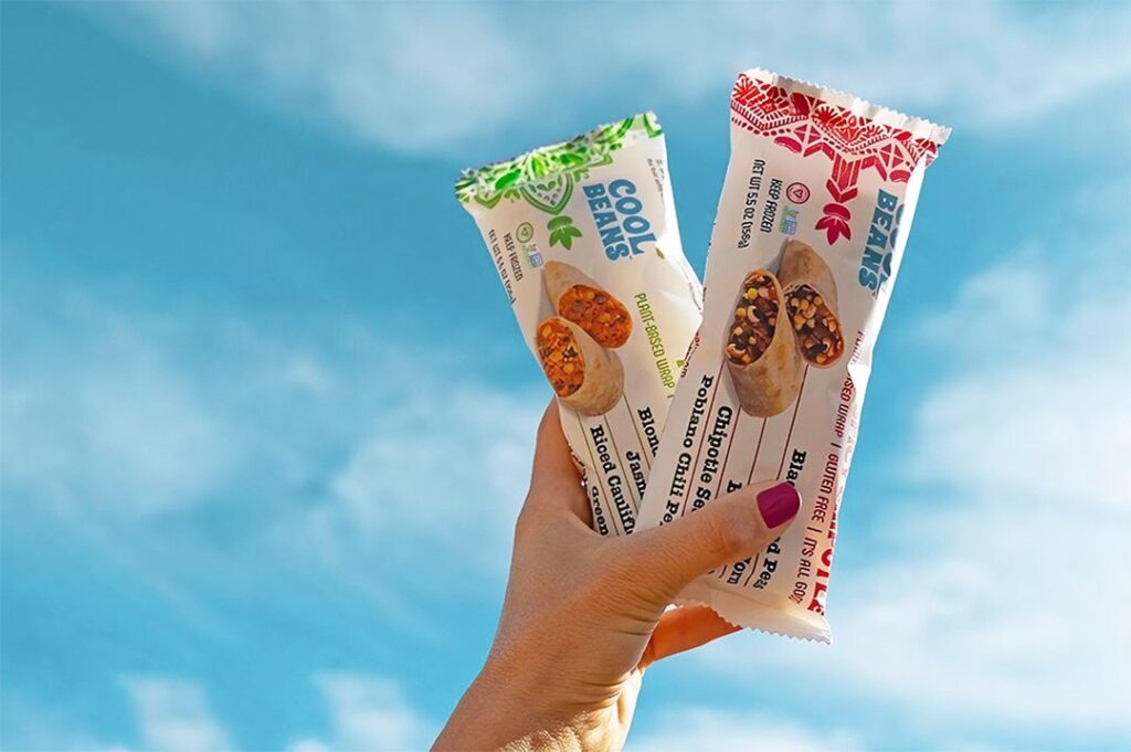 Photo shows a hand holding two vegan frozen burritos made by Cool Beans