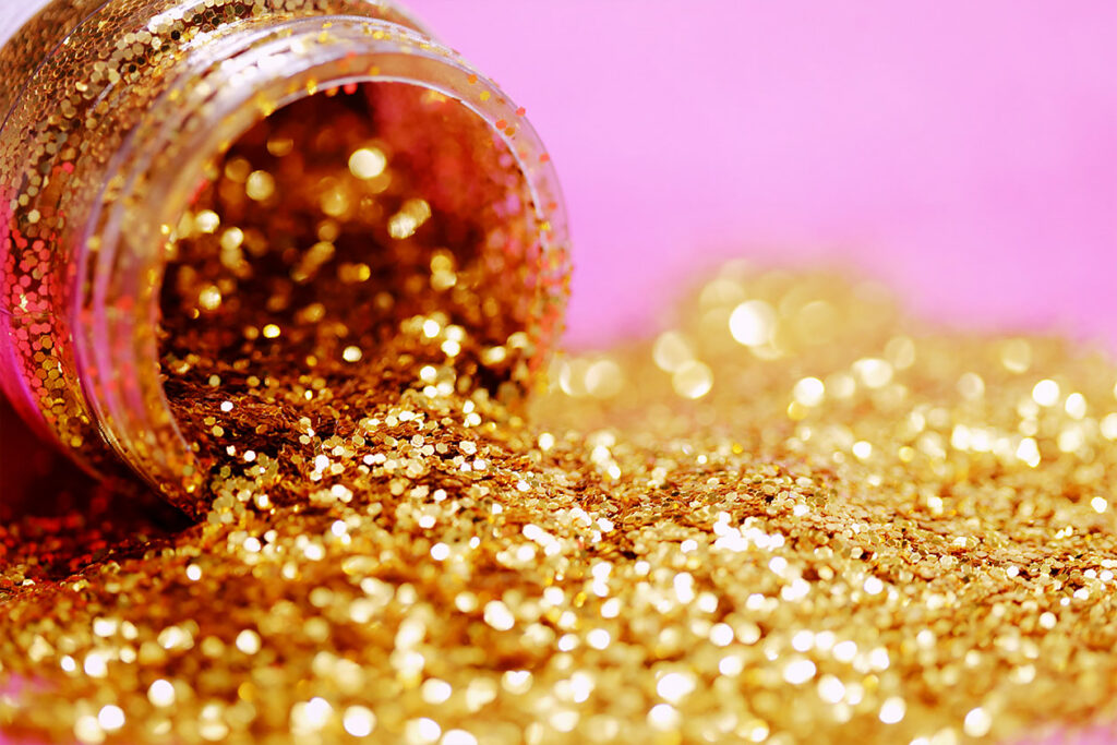 Photo shows a container of gold glitter tipping over on a pink background. Glitter particles are essential microplastics, and contribute to pollution.
