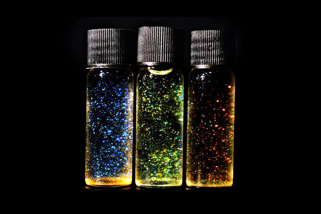 Photo shows three containers of glitter side by side on a black background, in blue, green, and orange, slightly lit from below. Can we still enjoy glitter if it contributes to plastic pollution?