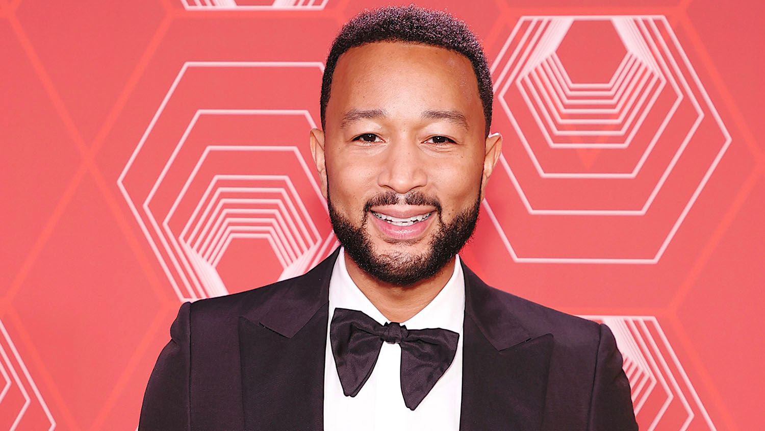 Photo shows John Legend in a tuxedo against a salmon pink background