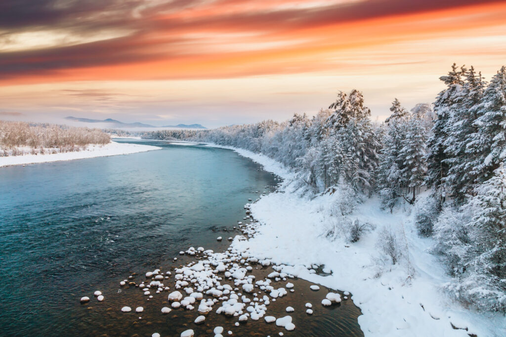 Photo shows a river flanked by snow against an orange-tinted sky.