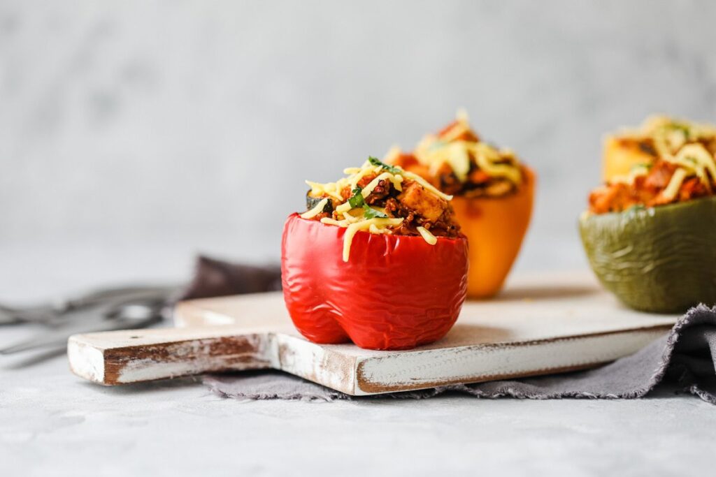 Photo shows three vegan stuffed peppers topped with dairy-free cheese shreds positioned on a cutting board